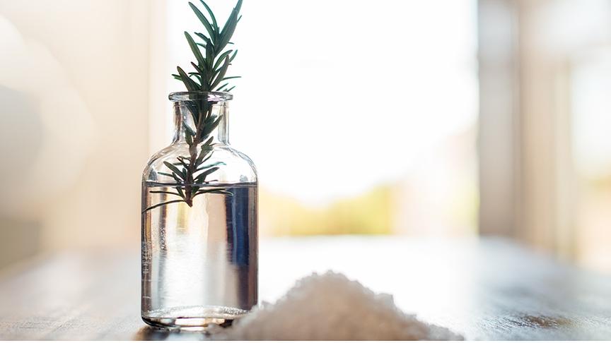 Ein kleines Wasserglas in dem ein einzelner Rosmarinzweig steckt, davor grobkörniges Salz in einem Haufen aufgeschüttet