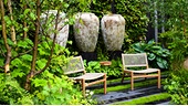 Schattige, sehr viel und dicht begrünte Gartenecke. In der Mitte stehen zwei tiefe, geflochtene Sessel mit einem kleinen Tisch. Im Hintergrund sind drei sehr große und hohe Töpfe.