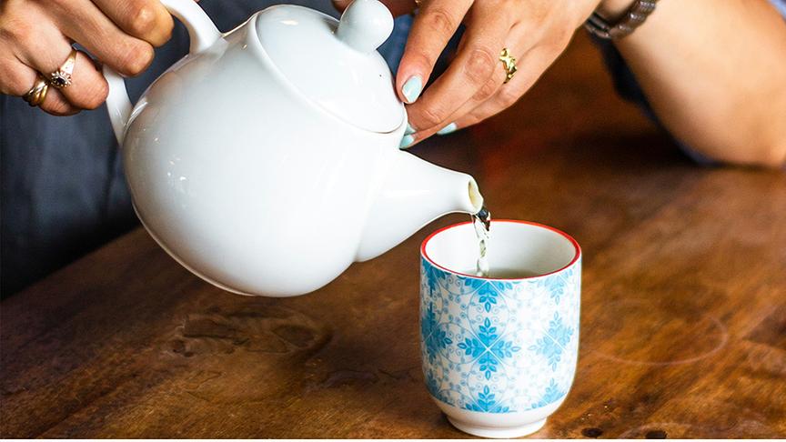 © Foto: Unsplash Eine Frau schenkt Tee von einer weißen Porzellanteekanne in einen blau gemusterten Becher.