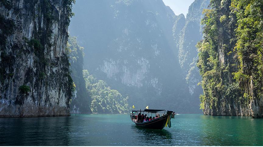 Ein traditionelles thailändisches Langschwanzboot mit Passagieren gleitet über das türkisblaue Wasser eines ruhigen Sees. Es ist von steilen, imposanten Kalksteinfelsen umgeben, die dicht mit üppigem Grün bewachsen sind und majestätisch aus dem Wasser ragen. Durch den leichten Dunst zwischen den Klippen bricht sanftes Sonnenlicht und verleiht der tropischen Landschaft eine friedliche, fast mystische Atmosphäre.