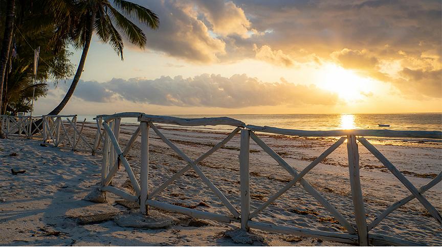 Ein stimmungsvoller Sonnenaufgang an einem tropischen Strand ein, bei dem das warme Licht der Sonne durch eine dichte Wolkendecke bricht. Im Vordergrund zieht sich ein rustikaler Holzzaun über den hellen Sand, während auf der linken Seite hohe Palmen sanft in das Bild ragen. Die Szene strahlt eine friedliche Ruhe aus und zeigt im Hintergrund das ruhige Meer unter einem weitläufigen Himmel.