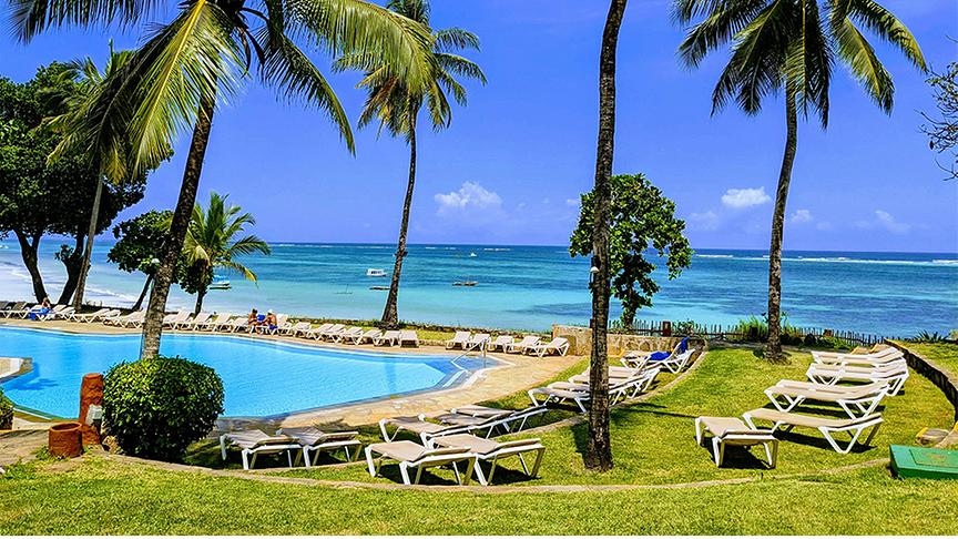 Eine sonnige Hotelanlage mit einem türkisfarbenen Pool im Vordergrund, der direkt an einen weißen Sandstrand und das azurblaue Meer grenzt. Üppige grüne Palmen ragen in den klaren blauen Himmel und spenden Schatten für die zahlreichen weißen Sonnenliegen, die auf den gepflegten Rasenflächen und am Beckenrand verteilt sind. Die tropische Kulisse strahlt eine entspannte Urlaubsatmosphäre aus und wird durch kleine Boote am Horizont des Indischen Ozeans ergänzt.