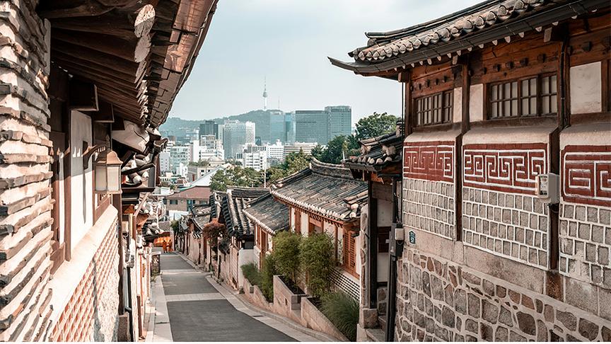 © Foto: Unsplash Gasse gesäumt von historischen, südkoreanischen Häusern mit Blick auf die Skyline Seouls im Hintergrund.