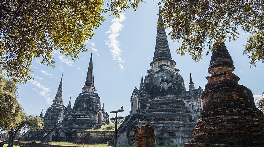 Eine Weitwinkelaufnahme zeigt die Ruinen einer antiken thailändischen Tempelanlage unter strahlend blauem Himmel mit leichten Schleierwolken. Im Vordergrund und an den Seiten rahmen grüne Baumkronen das Bild ein. Die historischen Backsteinbauten stehen auf einer weitläufigen Rasenfläche und zeugen von einer prachtvollen Vergangenheit.
