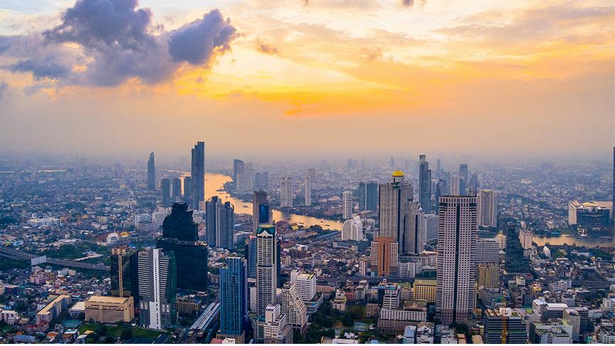 Weitläufige Skyline von Bangkok während der goldenen Stunde. Im Vordergrund und Zentrum ragen zahlreiche moderne Wolkenkratzer empor, darunter markante Gebäude wie der MahaNakhon mit seiner pixelartigen Architektur.Der Chao Phraya Fluss schlängelt sich im Hintergrund durch das dicht bebaute Stadtgebiet. Der Himmel ist in warme Gelb- und Orangetöne getaucht.