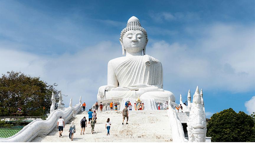 Beeindruckender Big Buddha von Phuket, eine riesige, weiße Statue aus Marmor, die majestätisch auf einem Hügel thront. Eine breite Treppe führt hinauf zur Figur, auf der zahlreiche Besucher unter einem strahlend blauen Himmel zu sehen sind. Die Szenerie wird von üppigem Grün eingerahmt.
