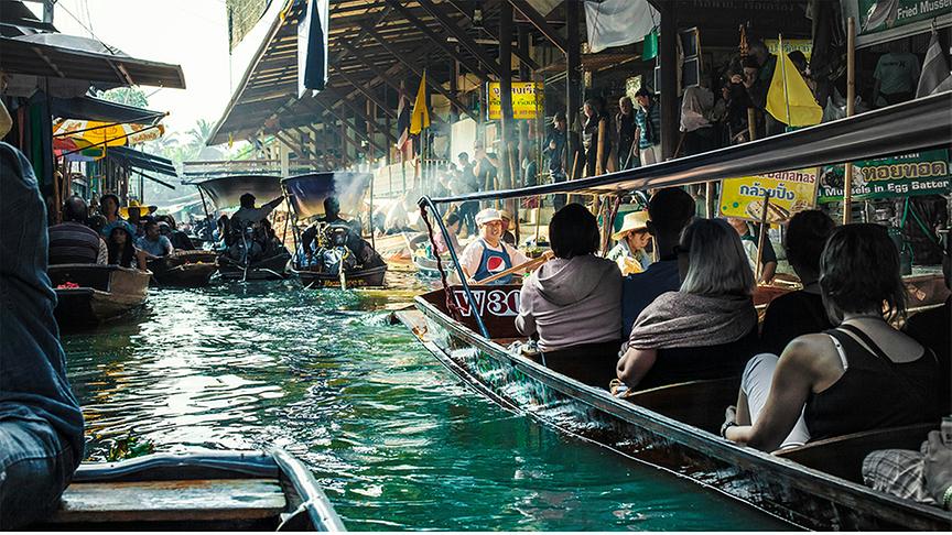 Belebter Damnoen Saduak Floating Market in Thailand, auf dem sich zahlreiche schmale Holzboote durch einen grünen Kanal schieben. Im Vordergrund blickt man von einem Boot aus auf weitere Passagiere und Händler, während im Hintergrund Sonnenstrahlen durch die hölzernen Marktstrukturen am Ufer fallen. Die Szene ist geprägt von einer dynamischen Atmosphäre mit Touristen, Einheimischen in traditionellen Hüten und bunten Sonnenschirmen entlang des Wasserwegs.