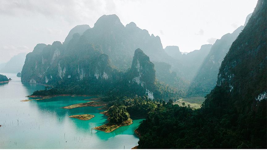 Eine weite Luftaufnahme zeigt die smaragdgrüne Wasserlandschaft des Cheow-Lan-Sees im Khao Sok Nationalpark, gesäumt von dicht bewaldeten Ufern. Im Hintergrund ragen markante, steile Kalksteinfelsen imposant in den dunstigen Himmel, während Sonnenstrahlen vereinzelt durch die Wolkendecke brechen. Die Szenerie vermittelt eine unberührte, mystische Atmosphäre dieser tropischen thailändischen Naturlandschaft.