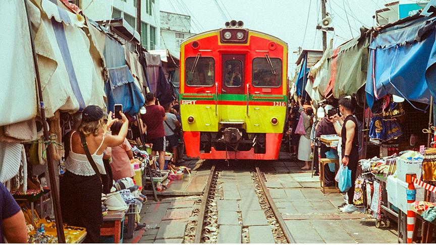 Ein farbenfroher rot-gelber Zug fährt unmittelbar durch die engen Gassen des Maeklong Railway Market in Thailand, während Touristen das Spektakel fotografieren. Die Marktstände auf beiden Seiten stehen so nah an den Schienen, dass ihre Markisen fast den Zug berühren. Die Szene ist belebt und zeigt das faszinierende Zusammenspiel zwischen täglichem Bahnbetrieb und traditionellem Markttreiben.