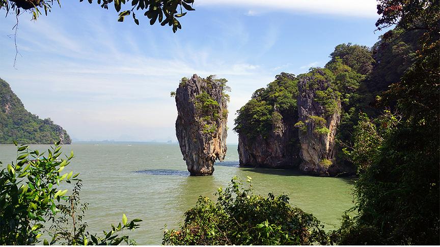 Eine malerische Fernaufnahme der berühmten James-Bond-Insel (Ko Tapu) in der Phang-Nga-Bucht, Thailand.Im Zentrum ragt eine markante, schmale Kalksteinfelsnadel steil aus dem grünlich schimmernden Meer empor. Sie ist im oberen Bereich mit etwas Vegetation bewachsen. Rechts daneben befinden sich größere, bewaldete Felsformationen. Eingerahmt wird die Szenerie durch dunkles Blattwerk im Vordergrund und an den Bildrändern, während der Himmel darüber hellblau und leicht bewölkt ist.