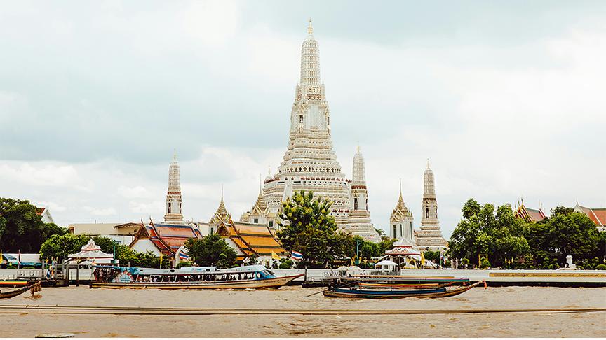 Weitwinkelaufnahme der weißen Tempelanlage Wat Arun in Bangkok. Im Vordergrund ein Fluss mit Booten, im Hintergrund ragt der Tempel mit mehreren spitzen Türmen empor.