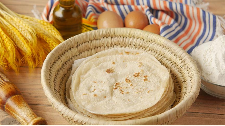 Geflochtener Korb mit Fladenbrot. Darum herum liegen Eier, Getreide, Mehl, Öl und ein gestreiftes Küchentuch.