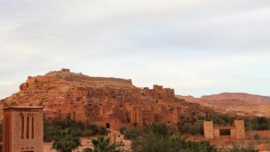 Historische Kasbah aus Lehm mit Palmen und Wüstenlandschaft im Hintergrund.