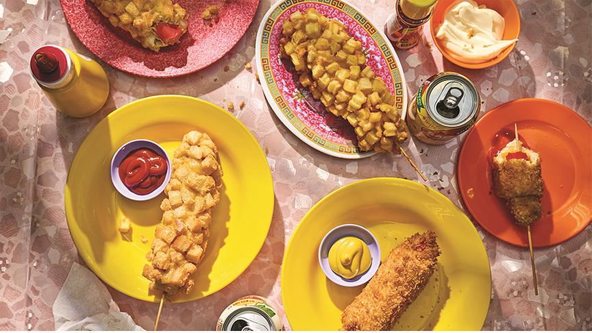 Dreierlei Hotdogs - auf bunten Tellern liegen unterschiedlich aussehende Korean Hotdogs mit einigen Soßen bzw. Dips und Getränkedosen.