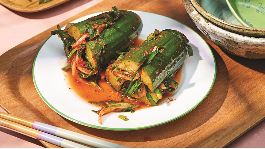 © © DK Verlag/ Daniel Herrmann-Zoll Gefüllter Gurken Kimchi. Die Gurken sind geviertelt, in der Mitte befindet sich das Kimchi. Der Teller ist auf einem holzfarbenen Tablett, daneben liegen Stäbchen.