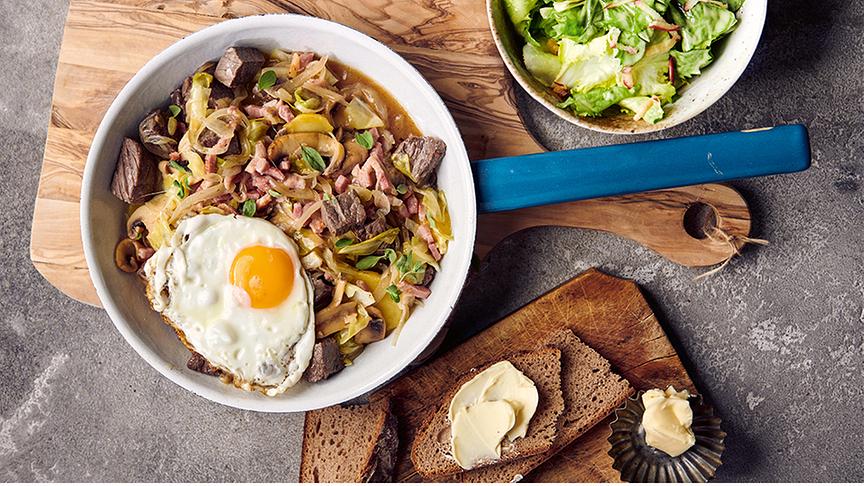 Eine Pfanne mit angebratenem Gemüse, Fleisch und einem Spiegelei. Daneben ein Brot mit Butter und Salat, schön angerichtet.