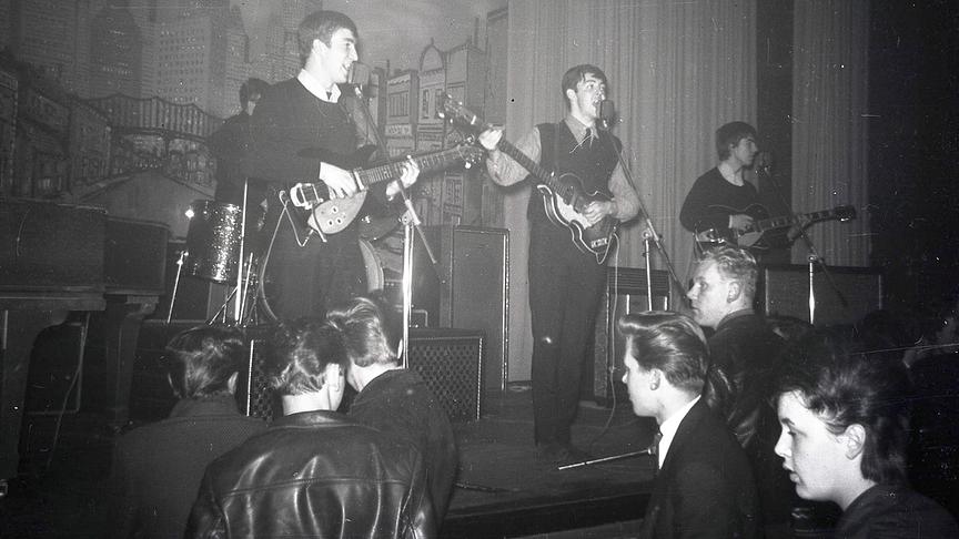 Eine aufgeblichene schwarz-weiß Aufnahme eines Auftritts der Beatles. Die vier Musiker stehen mit Instrumenten auf der Bühne vor einem jungen Publikum.