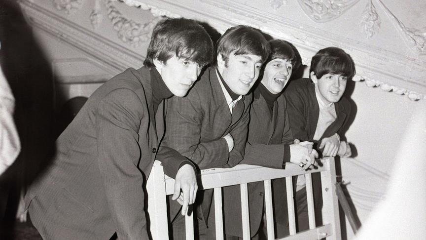 Die Beatles in Anzügen und charakteristischer Topffrisur an einem Geländer lehnend.
