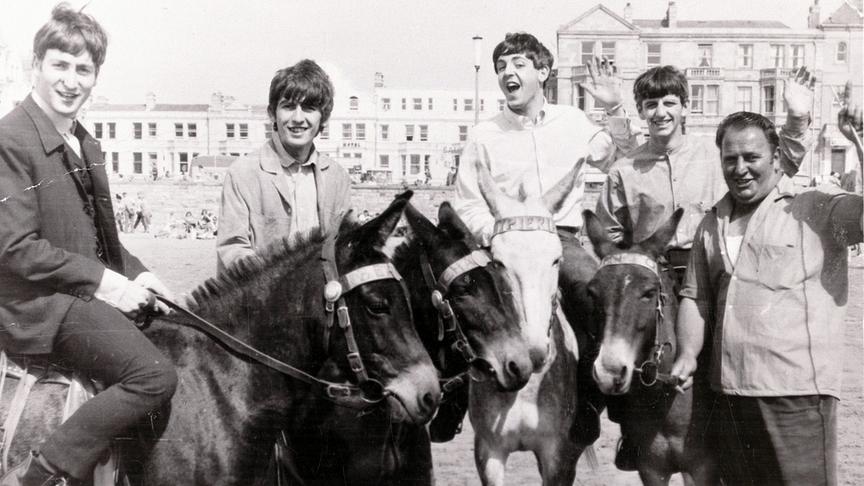 Die Beatles auf Eseln nebeneinander auf Esel sitzend.
