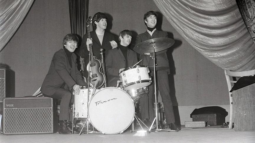 Schwarz-weiß Aufnahme der Beatles in Anzügen und mit Musikinstrumenten auf einer Bühne.