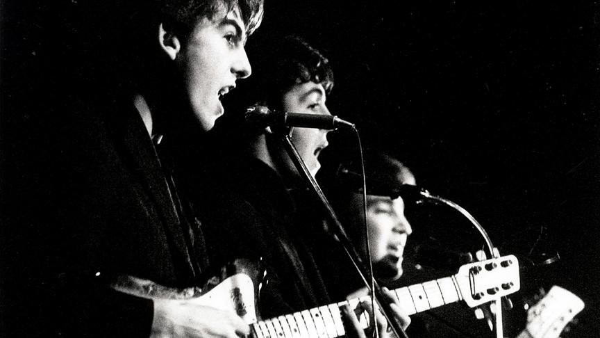 Schwarz-weiß Aufnahme der Beatles hinter Mikrofonen bei einem Auftritt.