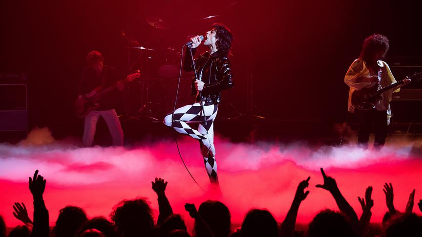 Mann mit schwarz-weiß gemusteter Hose singt in ein Mikrofon auf einer rot vernebelten Bühne vor Publikum