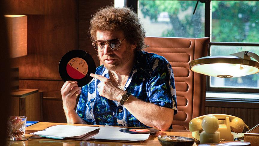 Mann sitzt am Schreibtisch, hält eine Schallplatte in der Hand und zeigt darauf