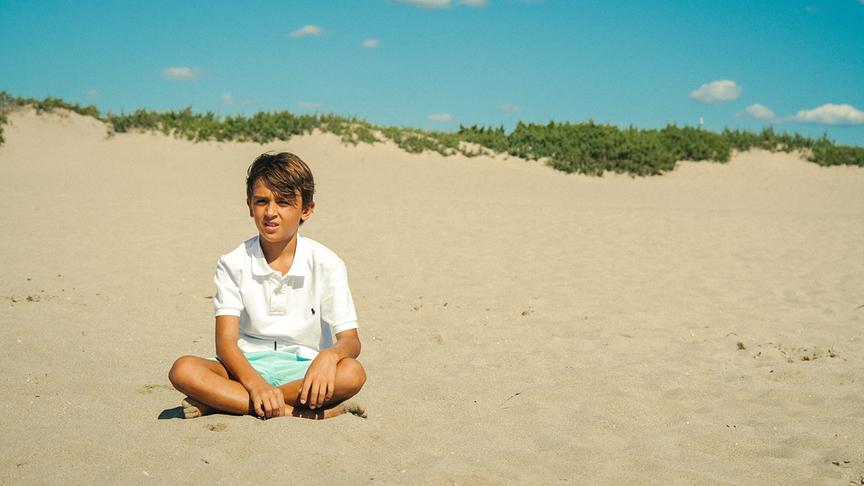 © Foto: ORF/ProSieben/LEONINE Studios Ein Junge sitzt im Schneidersitz am Sandstrand bei schönem Wetter.