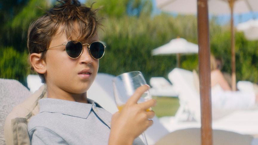 © Foto: ORF/ProSieben/LEONINE Studios Ein Junge mit Sonnenbrille liegt auf einer Sonnenliege und hält ein Weinglas mit Orangensaft in der Hand.
