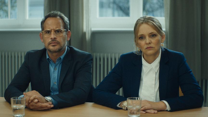 © Foto: ORF/ProSieben/LEONINE Studios Ein Mann und eine Frau im Anzug sitzen mit verschränkten Händen an einem Tisch und blicken ernst.