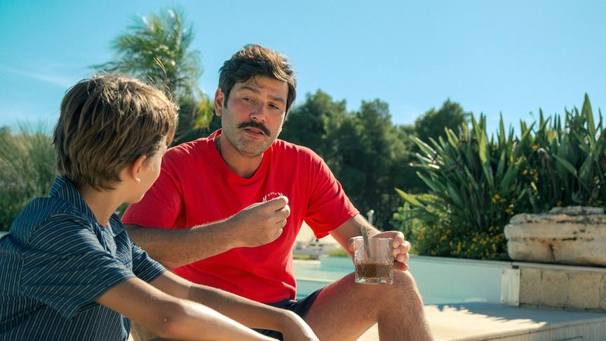 © Foto: ORF/ProSieben/LEONINE Studios Ein Mann mit Schnauzbart, rotem Tshirt und einem Getränkeglas in der Hand spricht zu einem Jungen, der neben ihm sitzt.
