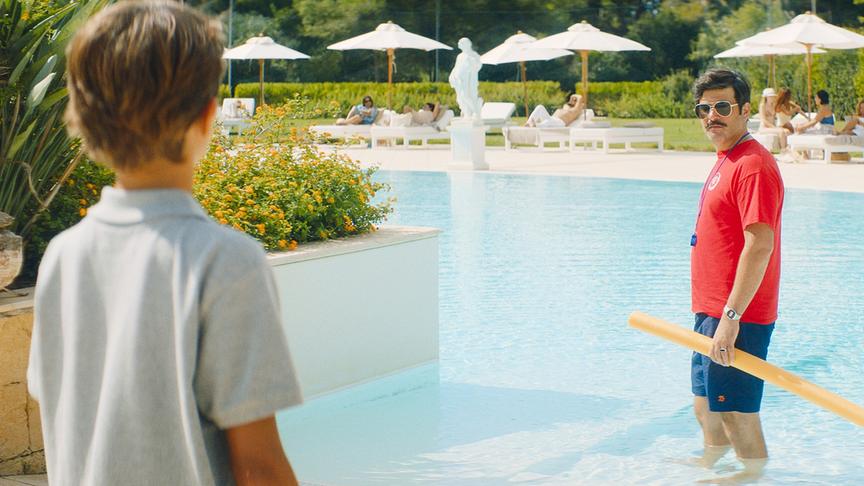© Foto: ORF/ProSieben/LEONINE Studios Ein Mann mit rotem Oberteil, kurzer Hose und Sonnenbrille steht knietief in einem Pool, hält eine Poolnudel in der Hand und blickt einen Jungen an, der am Rande der Pools steht. Im Hintergrund, auf der anderen Seite des Pools liegen mehrere Badegäste auf weißen Sonnenliegen unter weißen Sonnenschirmen.