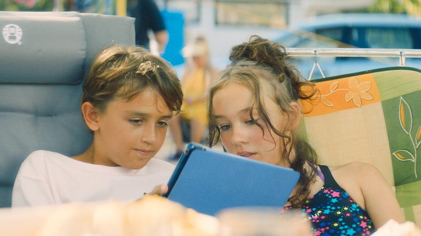© Foto: ORF/ProSieben/LEONINE Studios Ein Mädchen und ein Junge sitzen auf Campingstühlen und schauen gemeinsam in ein Tablet.