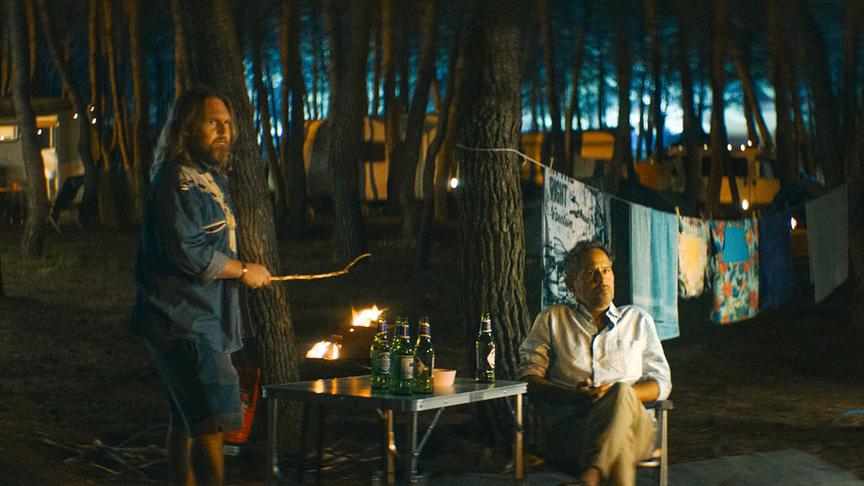 © Foto: ORF/ProSieben/LEONINE Studios Szene auf einem Campingplatz in der Dunkelheit. Zwei Männer, einer stehend und einer sitzend, an einem Campingtisch. Ein Mann hält einen Stock über offenes Feuer. Beide blicken in eine Richtung. Im Hintergrund hängen mehrere Handtücher an einer Wäscheleine und zwischen den Bäumen stehen Campingmobile.