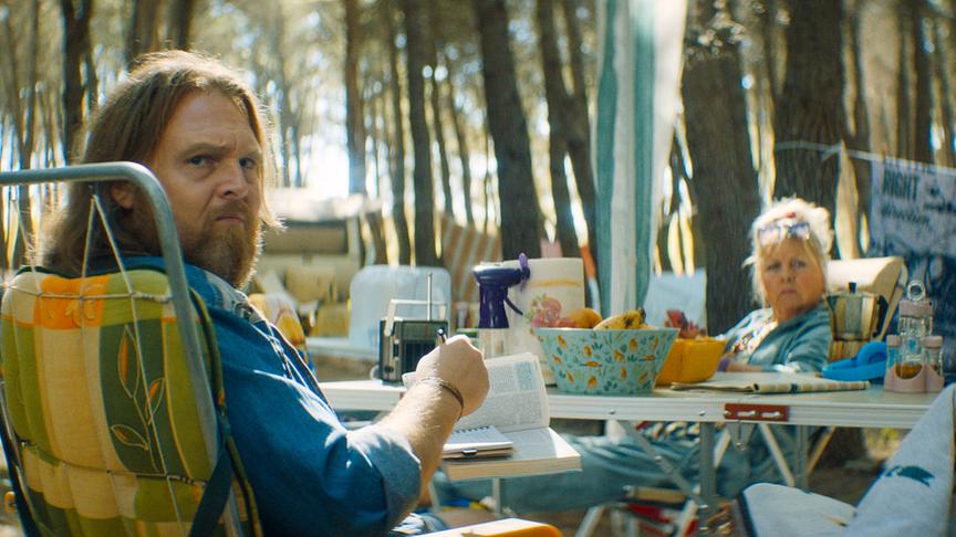 © Foto: ORF/ProSieben/LEONINE Studios Ein Mann mit langen blonden Haaren und mittellangem Bart und eine blonde Frau mit Hochsteckfrisur in legerer Kleidung sitzen an einem vollbepackten Campingtisch umgeben von weiterem Campingequipment. Sie blicken beide in Richtung der Kamera.