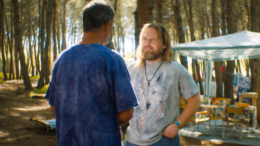 © Foto: ORF/ProSieben/LEONINE Studios Zwei Männer in legerer Kleidung stehen sich im Wald auf einem Campingplatz gegenüber. Der Mann, dessen Gesicht zur Kamera gewandt ist, trägt lange blonge Haare und einen mittellangen Bart.