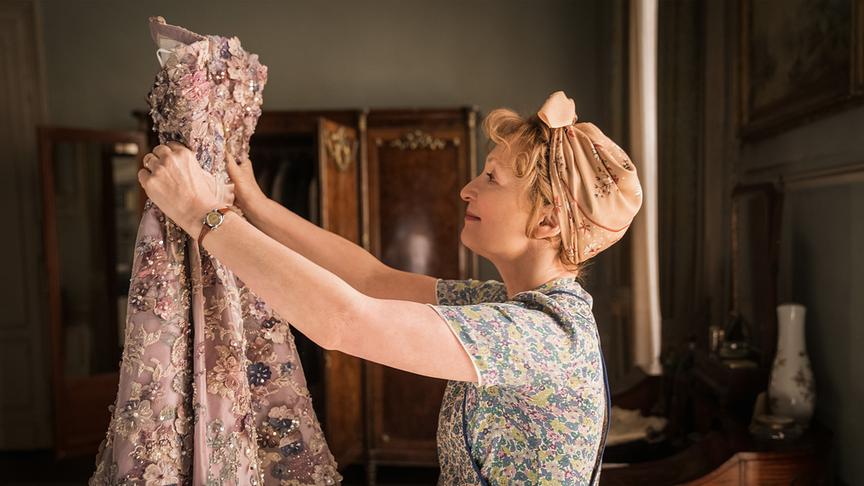 Eine Frau mit Tuch in den Haaren hält ein aufweniges Abendkleid vor sich in die Höhe.