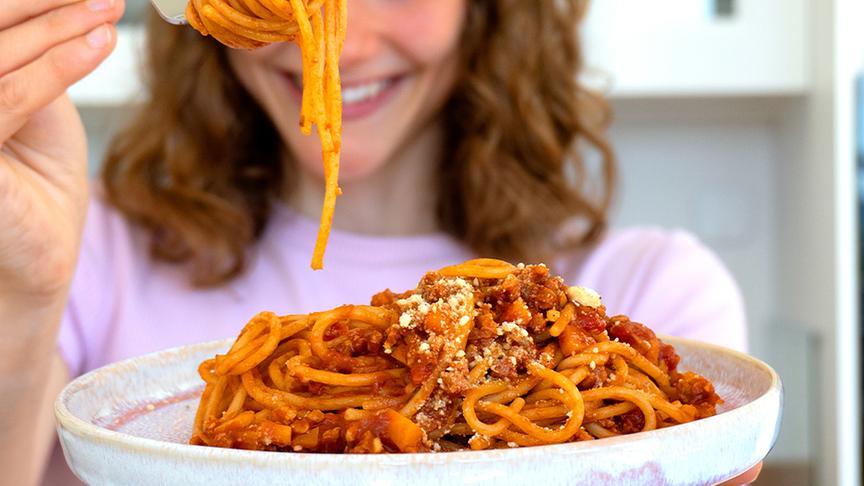 Maya Leinenbach hält einen Teller mit Spaghetti Bolognese vor sich. In der rechten Hand hält eine Gabel auf der ein paar Spaghetti aufgerollt sind.