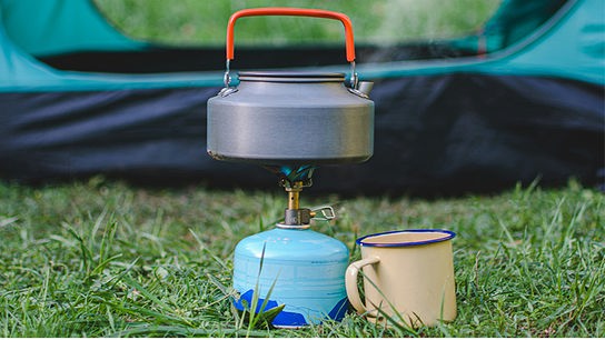 Ein Kessel auf einem kleinen Campingkocher, daneben eine Tasse. Im Hintergrund ein Zelt.