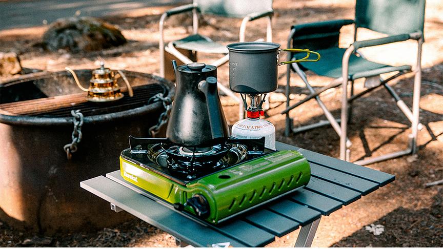 Camping-Kochausrüstung mit Campingkocher, Wasserkessel, Tisch, Campingstühlen und Feuerstelle.