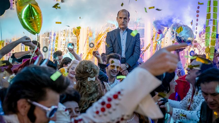 Eine große, bunte Feier mit vielen verkleideten Personen und buntem Rauch. Im Hintergrund ein nicht dazupassender Mann in einem normalen, grauen Anzug.