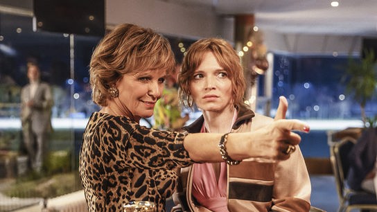 © Foto: ORF/ProSieben/Hellinger/Doll Filmproduktion GmbH/Warner Bros. Zwei Fauen stehen dicht nebeneinande an de Bar. Die ältere Fau trägt ein Leopardenoberteil, auffälligen Schmuck und zeigt mit Zeigefinger und Daumen wie mit einer Pistole auf etwas außerhalb des Bildes. Die jüngere Frau ist legerer gekleidet und blickt in die Richtung, in die die andere Frau zeigt. Vo den Frauen auf der Bar steht ein gefülltes Glas und eine kleine Handtasche.