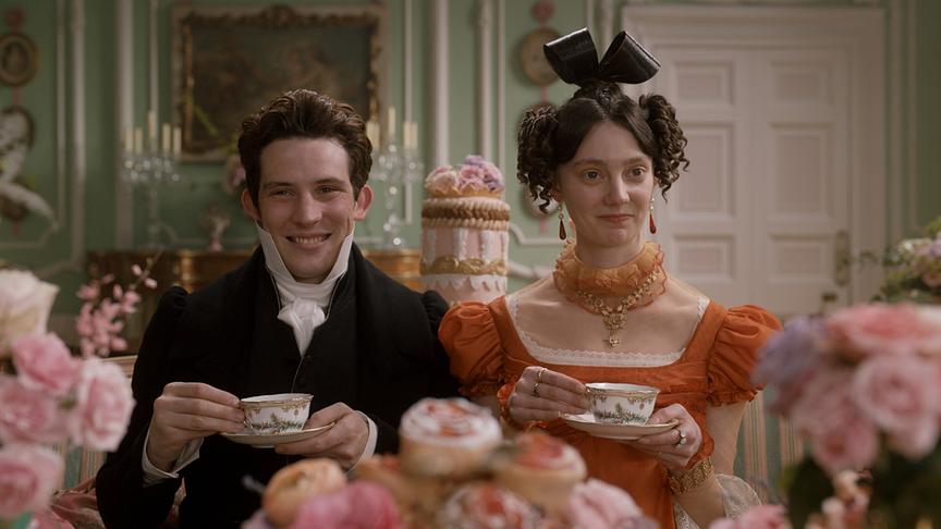 Ein Mann und eine Frau sitzen lächeln mit Teetasse nebeneinander, um sie herum Desserts und Rosen.