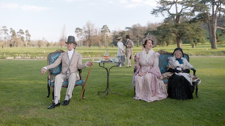 Drei Personen sitzen auf einem Rasen auf einem Sessel und einem Sofa, das eher für drinnen bestimmt zu sein scheint, im Hintergrund stehen zwei weitere Personen.