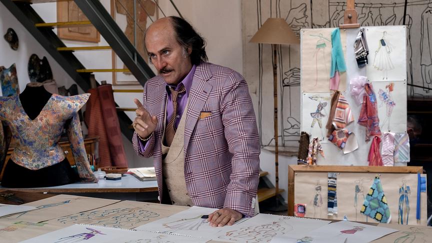 Ein Mann in einem Atelien mit Stoffmustern und -schnitten sowie Entwürfen steht