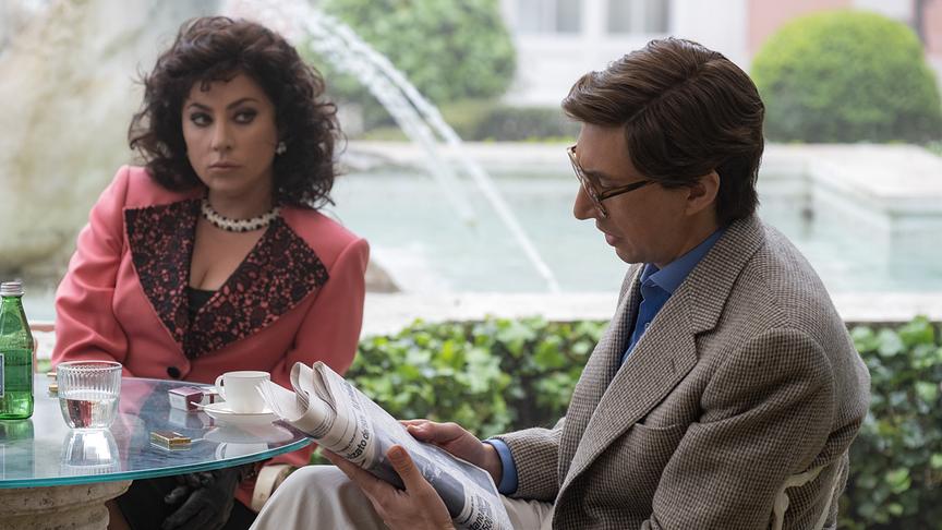 Eine Frau und ein Mann an einem Tisch sitzend, die Frau blickt zum Mann, während dieser Zeitung liest