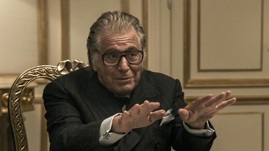 Ein Mann auf einem goldenen Stuhl sitzend gestikuliert mit beiden Armen ausgestreckt und Handflächen nach unten gerichtet