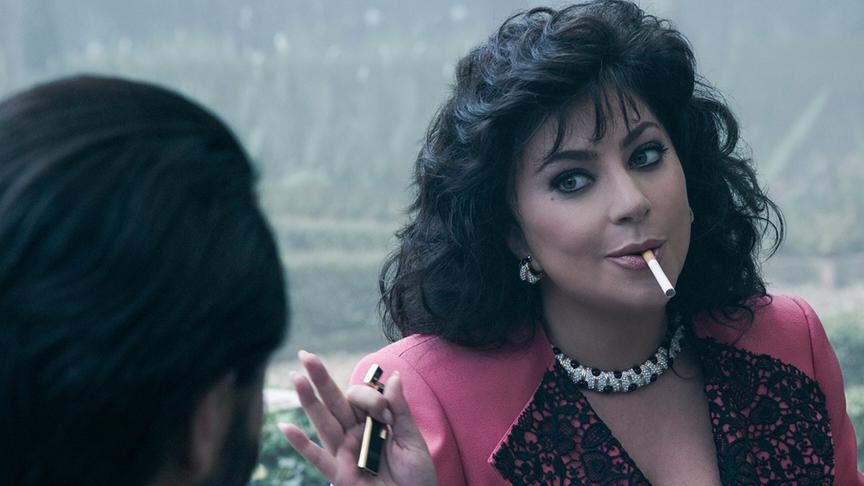 Eine Frau mit dunklen Locken, erhobenen Handflächen und Zigarette im Mund sitzt einem Mann gegenüber