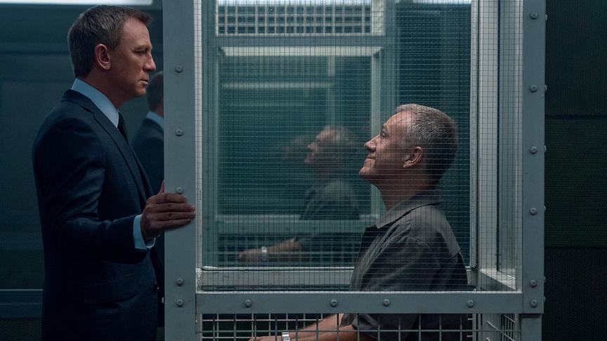 Ein Mann steht dicht vor einem stahlverstärktem Glaskasten, in dem ein Mann mit Handschellen sitzt.