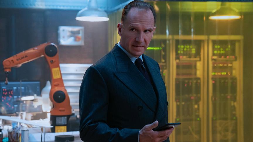 Ein Mann in Anzug und mit Handy in der Hand vor technischen Geräten und einem Roboterarm.